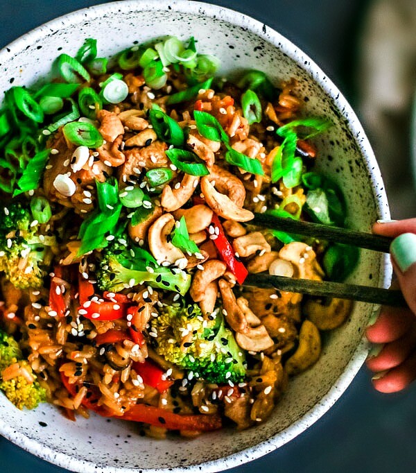Hands holding a bowl filled with rice, chicken, vegetables, and cashews.