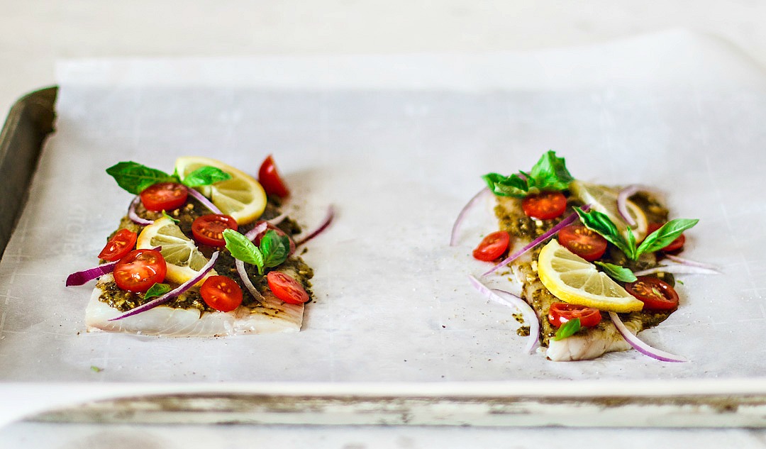 Side shot of flounder fillets sitting on parchment paper topped with bright ingredients.