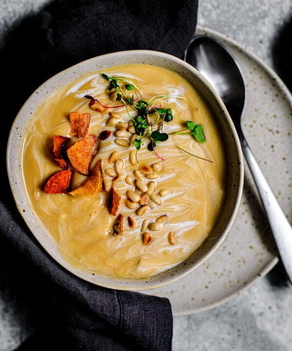 Bowl of roasted garlic and parsnip soup garnished with toasted pine nuts, root chips, and microgreens.