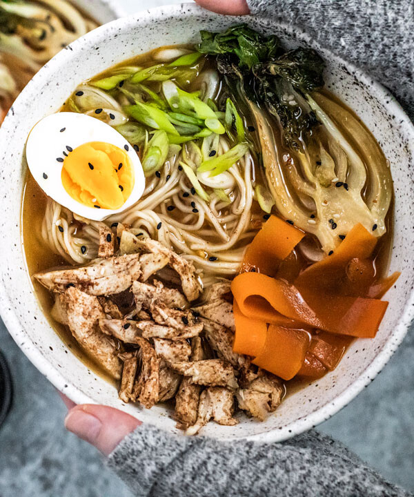 Hands holding up five spice chicken Ramen bowl topped with an egg and vegetables.