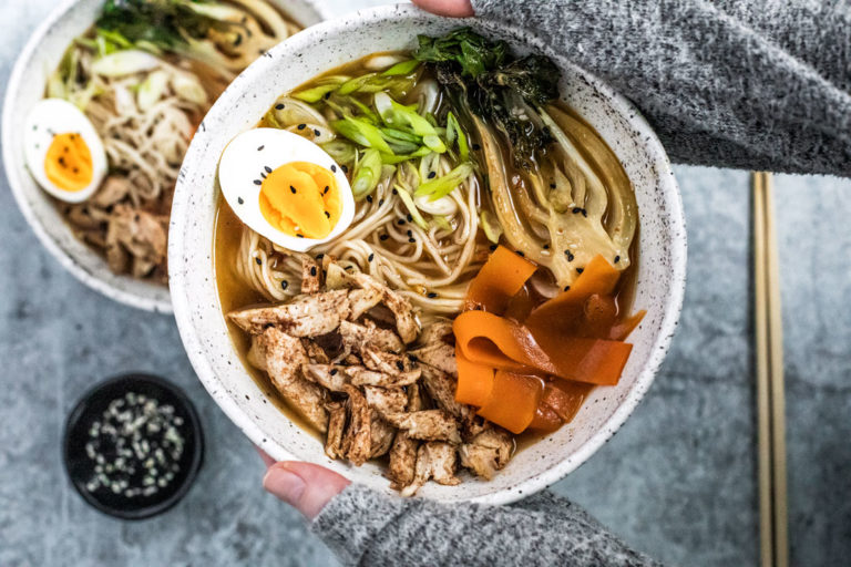 Hands holding up five spice chicken Ramen bowl topped with an egg and vegetables.