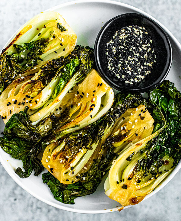 Round white plate stacked with bright green seared baby bok choy with a black pinch bowl full of black and white sesame seeds.