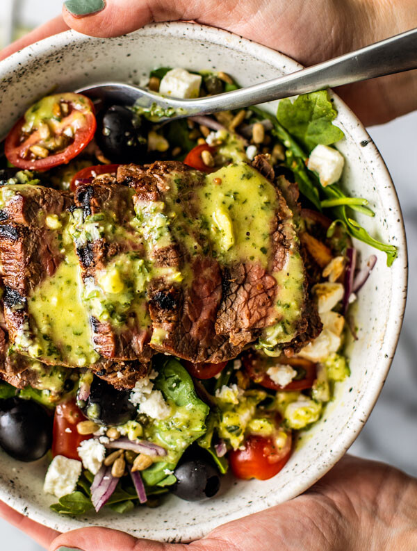 Hands holding up a bowl of Greek Steak Salad drizzled with herb and garlic vinaigrette.