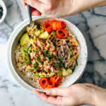 Hands holding a big bowl of rice, mackerel, cucumber, avocado, and carrots.