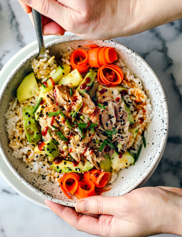 Hands holding a big bowl of rice, mackerel, cucumber, avocado, and carrots.