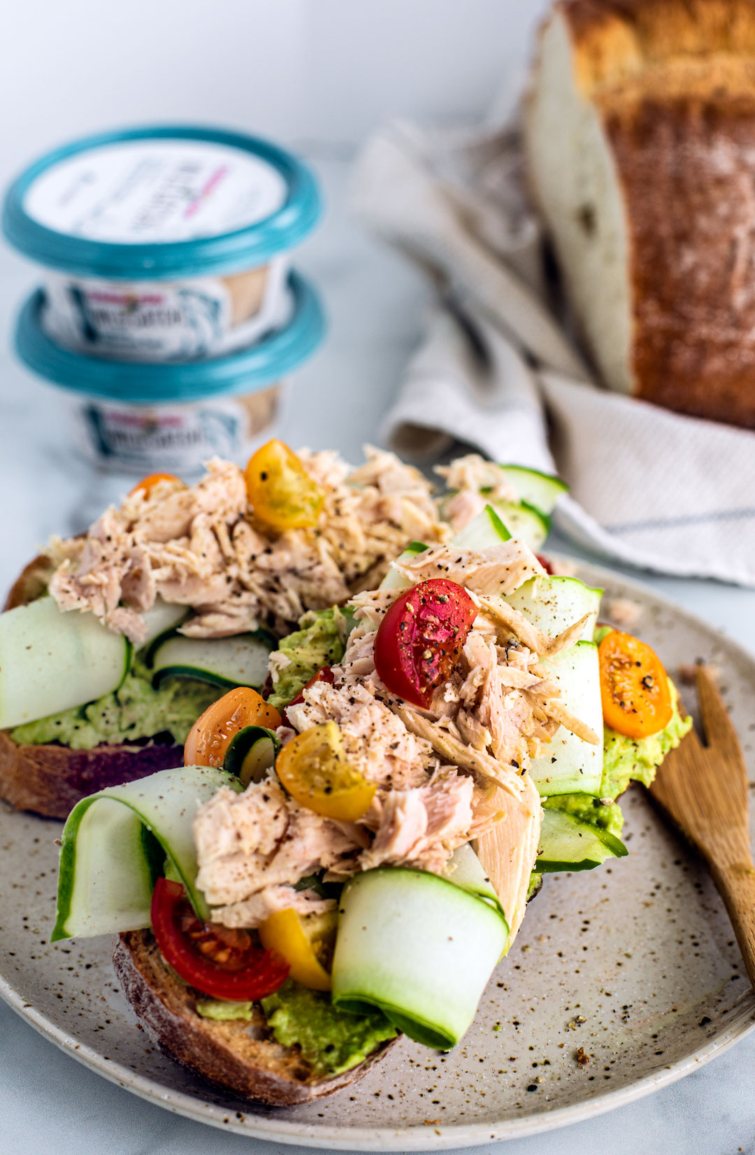 Two slices of toast topped with smashed avocado, ribboned cucumber, tomatoes, and flaked tuna.