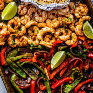 Sheet pan covered with peppers, onions and shrimp with some lime wedges and soft shell tortillas on the side.