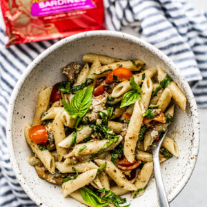 Overhead shot of a bowl of pesto pasta with tomatoes and fresh herbs and King Oscar can in the background.