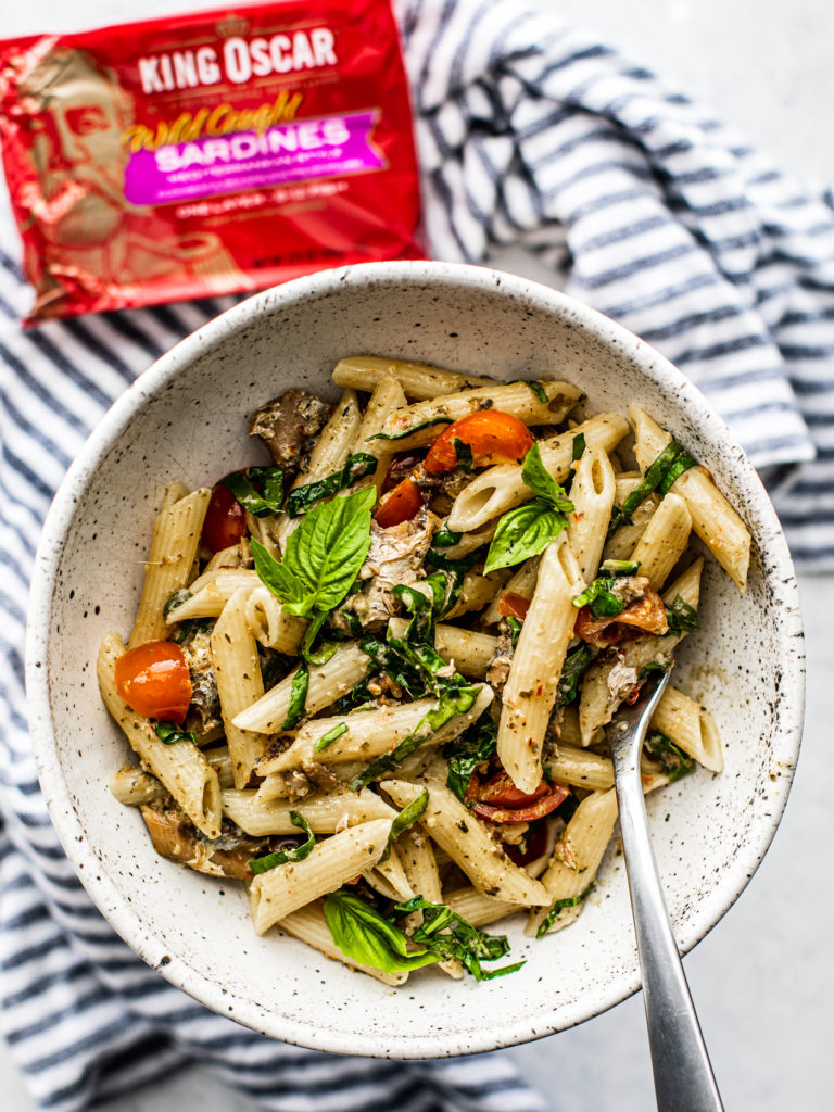 Overhead shot of a bowl of pesto pasta with tomatoes and fresh herbs and King Oscar can in the background.