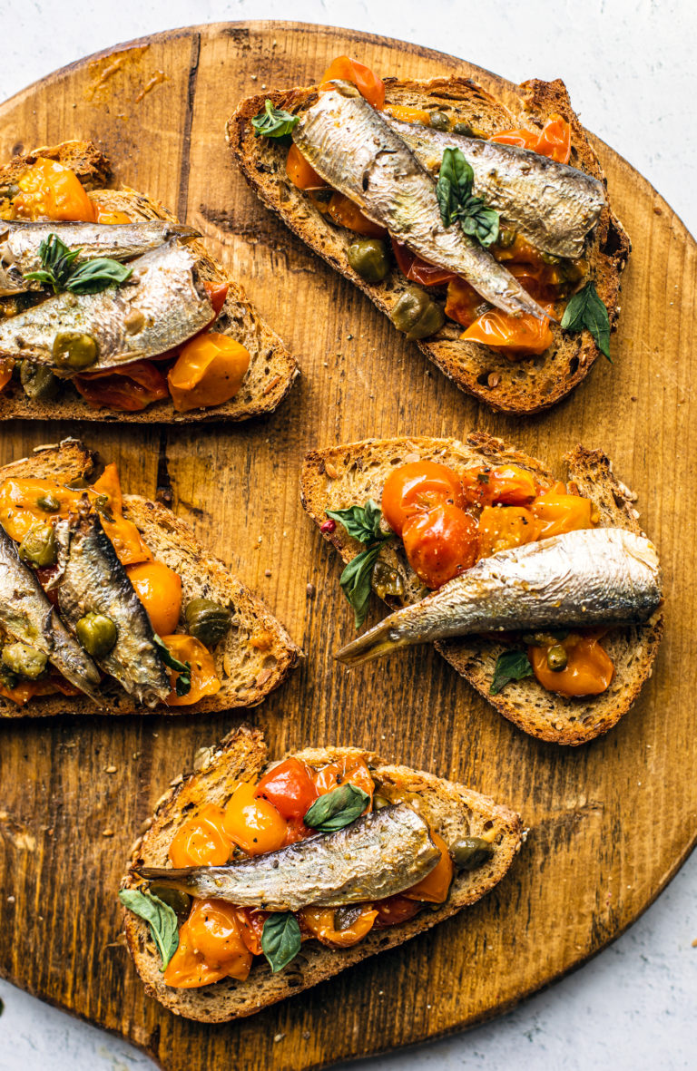 Wooden serving board with toasts topped with tomato confit and sardines.