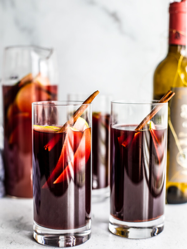 Two tall glasses of red sangria with sliced green apples and cinnamon sticks in it.