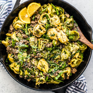 Cast iron skillet with tortellini and sausage, with a serving spoon in it.