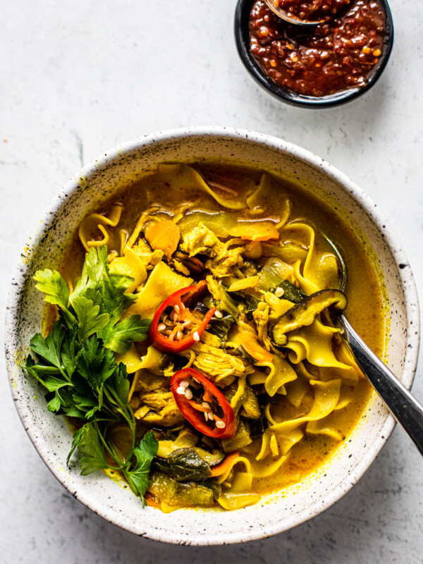 Bowl of curry chicken noodle soup garnished with fresh parsley and sliced chiles, and a small bowl of hot sauce next to it.