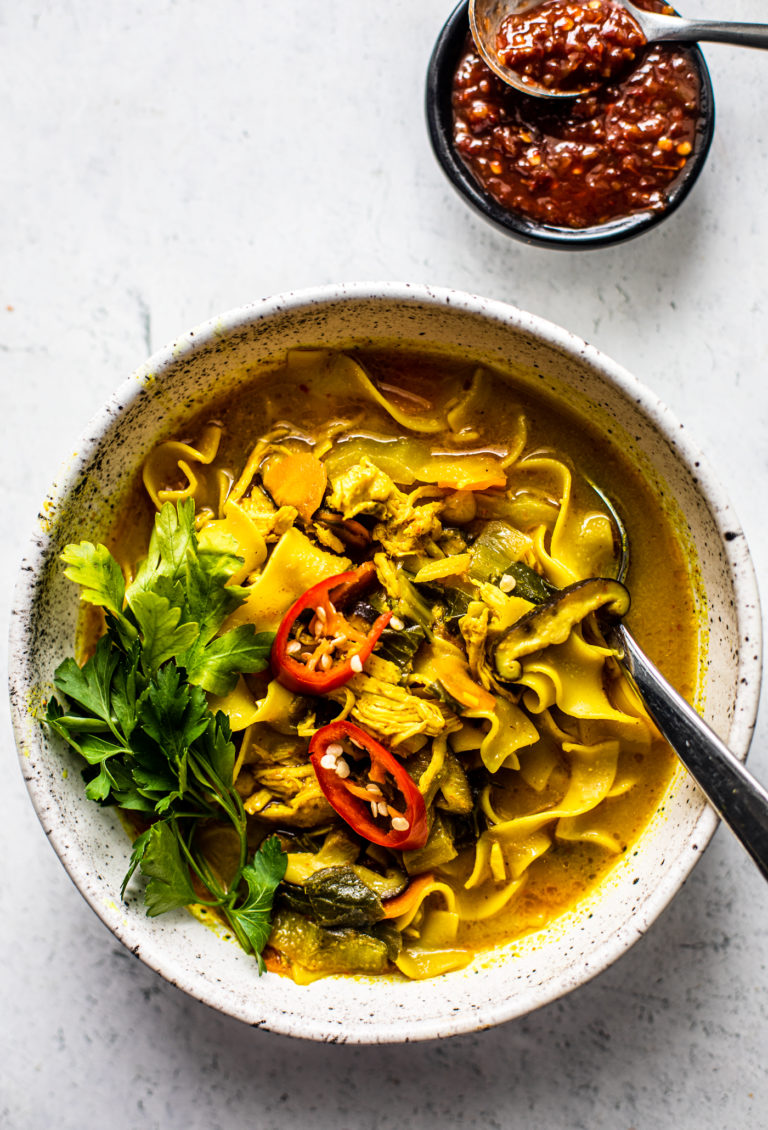 Bowl of curry chicken noodle soup garnished with fresh parsley and sliced chiles, and a small bowl of hot sauce next to it.