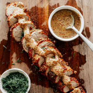 Sliced pork tenderloin on a wooden board drizzled with mustard sauce.