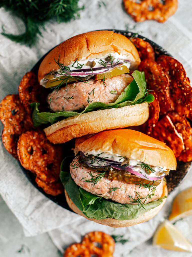 Two salmon burgers in a serving dish stacked with lettuce, pickles, onions, and mayo, with side of pretzels.