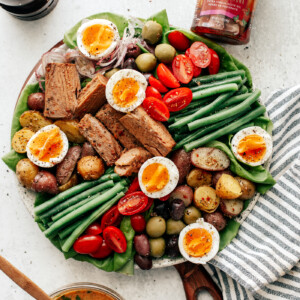 Colorful platter of Niçoise Salad with fresh veggie and soft-boiled eggs. Striped dish towel off to the side with bowl of vinaigrette and jar of tuna.