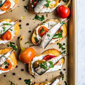 Golden sheet pan with crostini on it, which are topped with goat cheese, cherry peppers, and sardines.