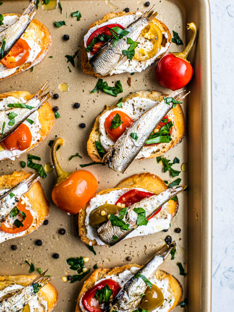 Golden sheet pan with crostini on it, which are topped with goat cheese, cherry peppers, and sardines.