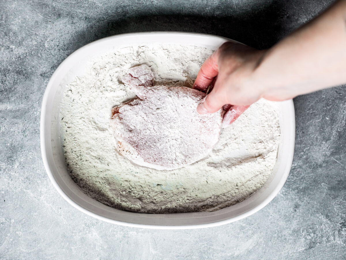 Hand taking floured pork chop out of dish of flour.