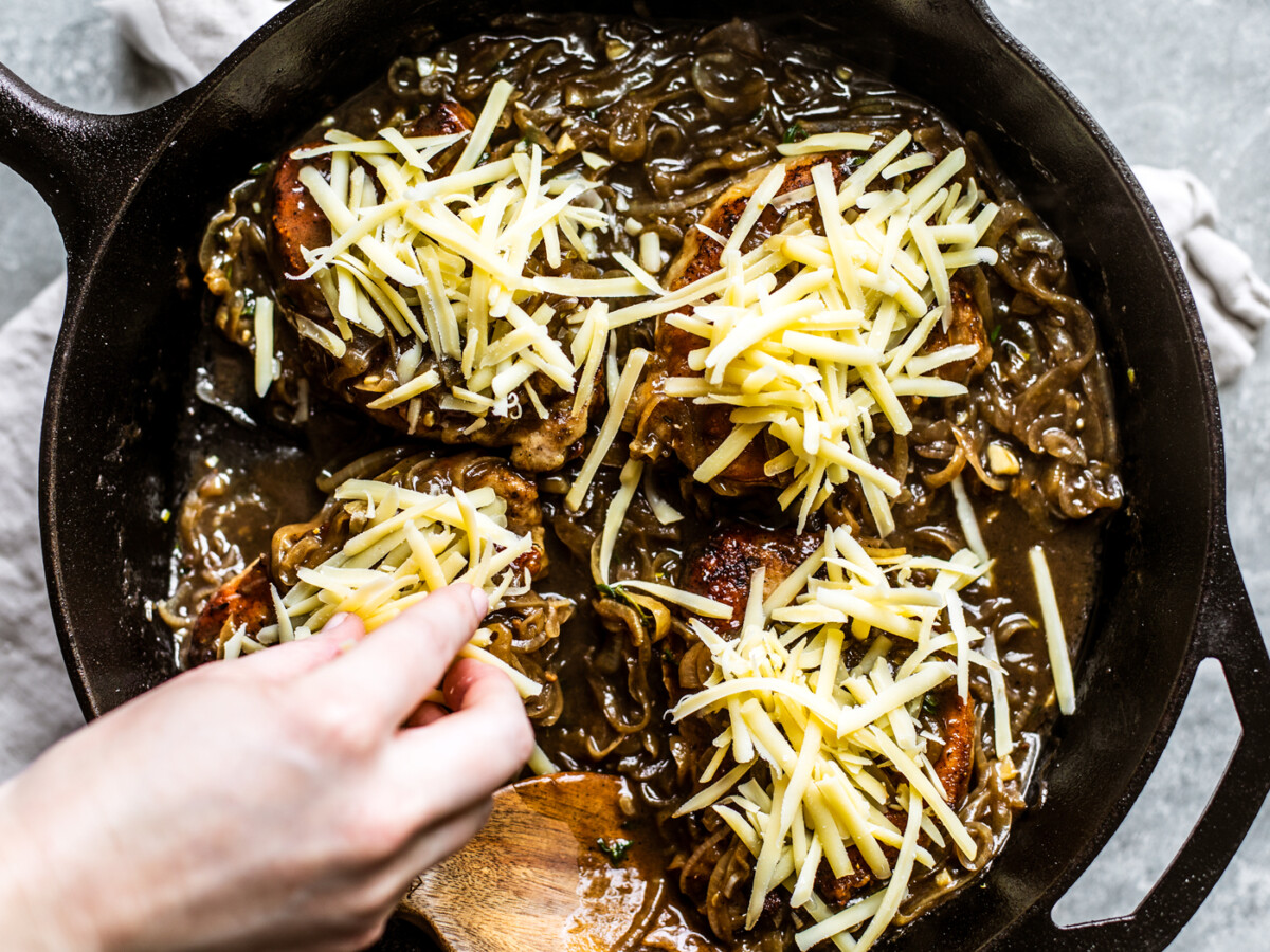 Grated cheese being put on top of pork chops in skillet.