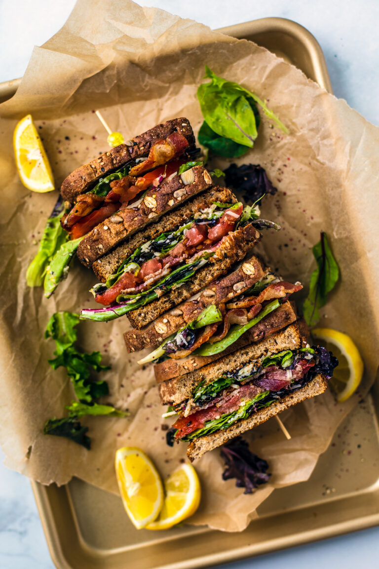 Four BLT sandwich halves on a serving platter with parchment, lemon wedges on the side.