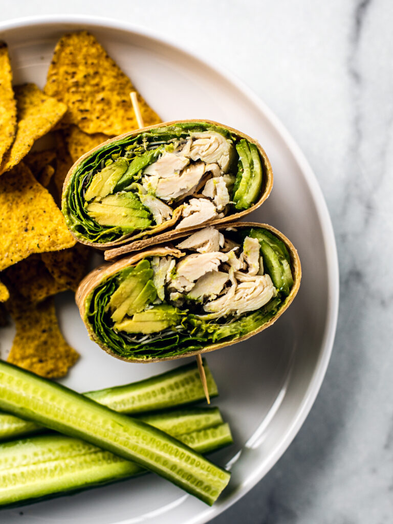 Close up of green goddess wrap on a white plate with a side of chips and cucumber spears.