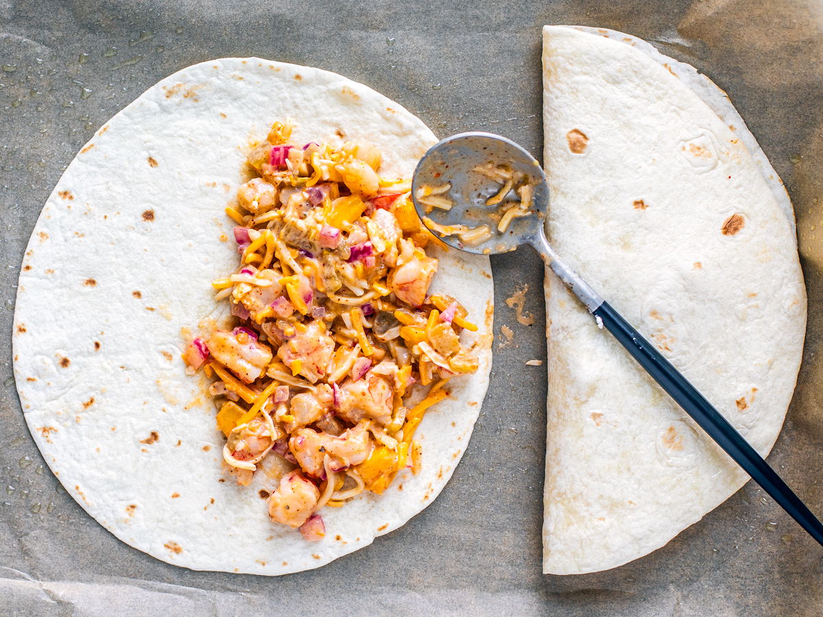 Sheet pan with on folded tortilla, and one being filled with shrimp quesadilla mix with a spoon to the side.