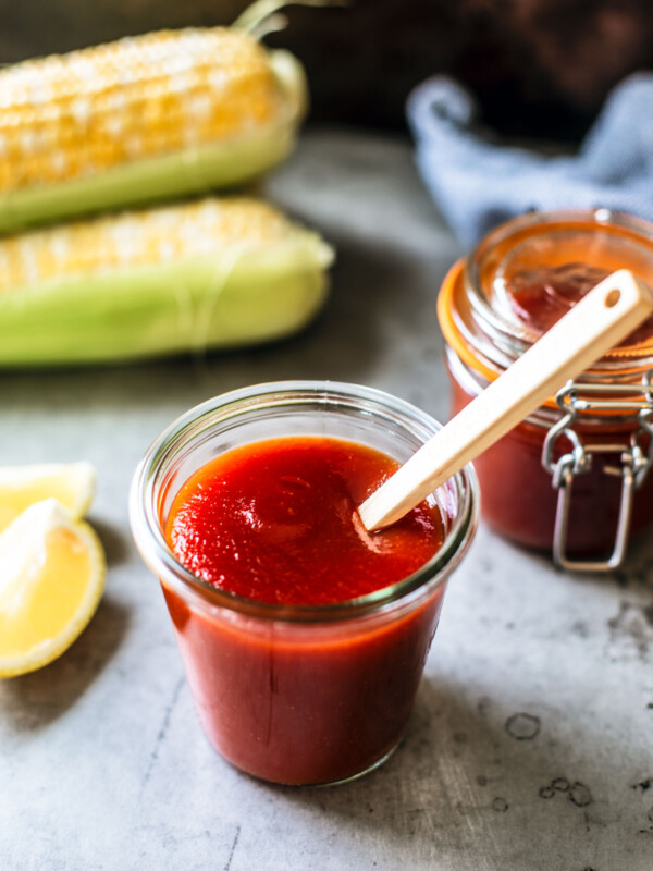 Jar of homemade BBQ sauce with a spoon in it with lemons and corn on the cob in the background.