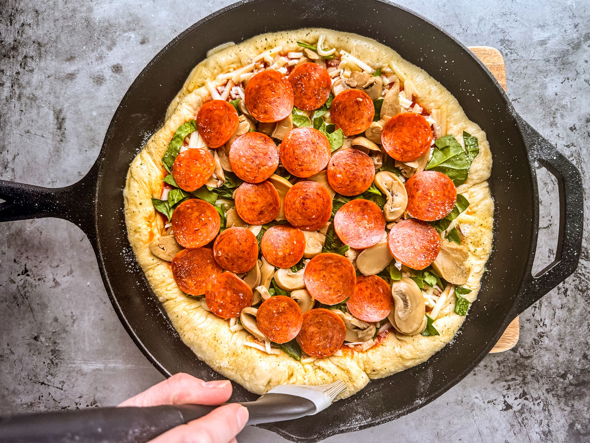 Toppings on skillet pizza with oil being brushed over the crust.