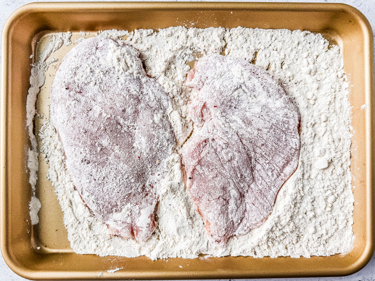 Chicken cutlets dredged in flour in a baking tray.