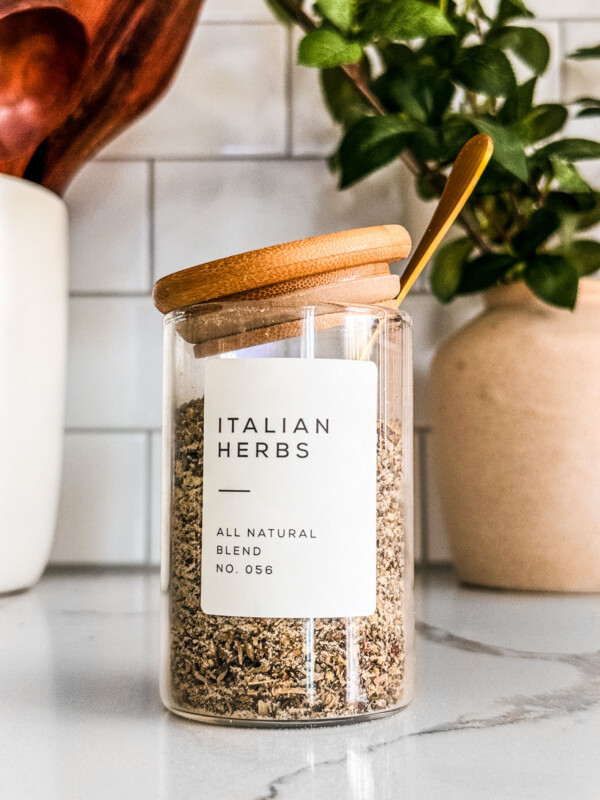 Glass jar of Italian seasoning on a white marble countertop.