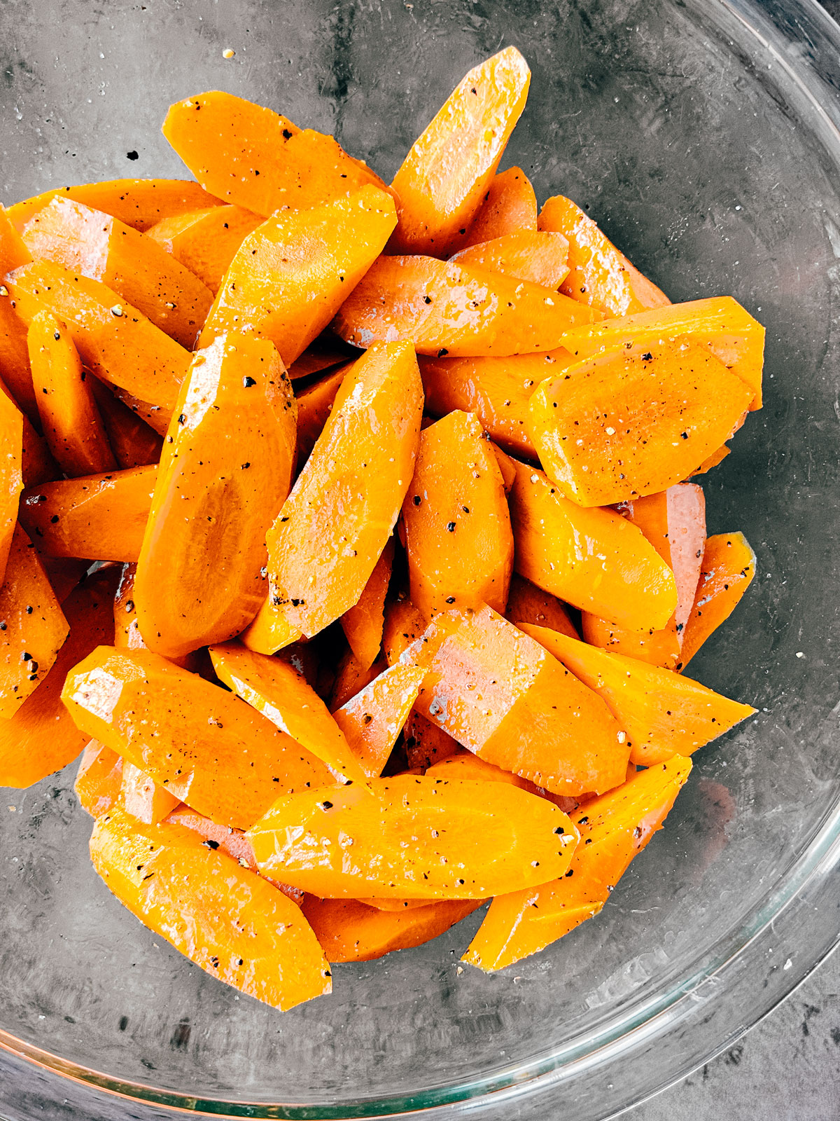 Mixing bowl with carrots tossed in olive oil, salt, and pepper.