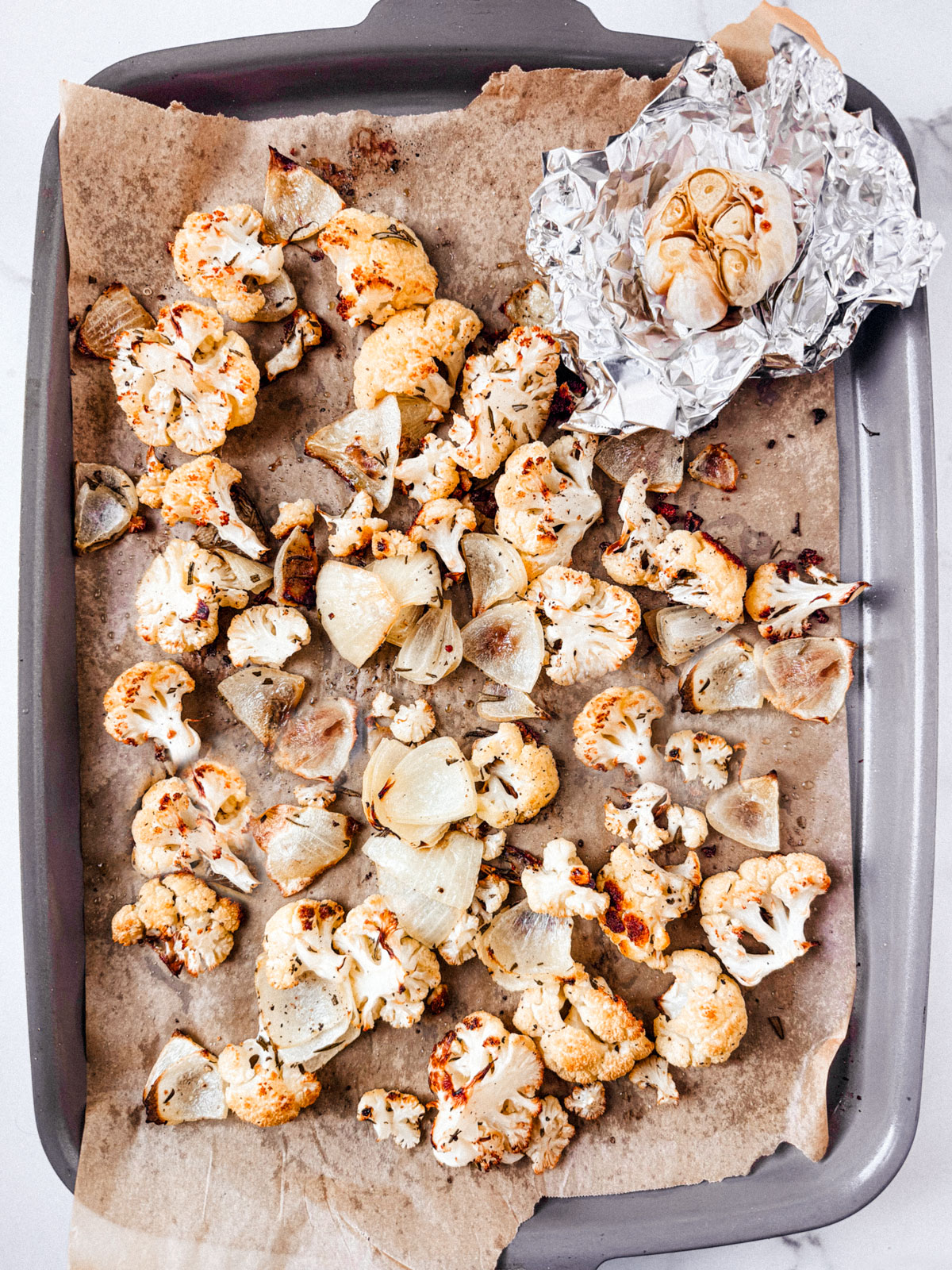 Roasted cauliflower and onions on a baking sheet with a packet of roasted garlic.