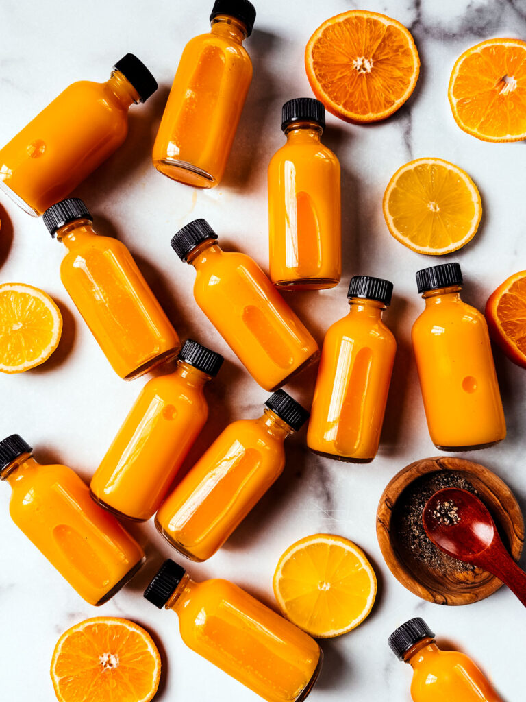 Turmeric ginger shots spread out over a white marble background with lemon and orange slices.